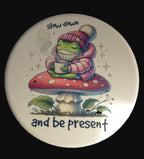 Coaster with a frog wearing a hat and scarf, sitting on a mushroom, holding a cup with text 'slow down and be present' on a purple surface.