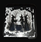 Decorative coaster with black and white illustration of three witches by a cauldron on a wooden surface.