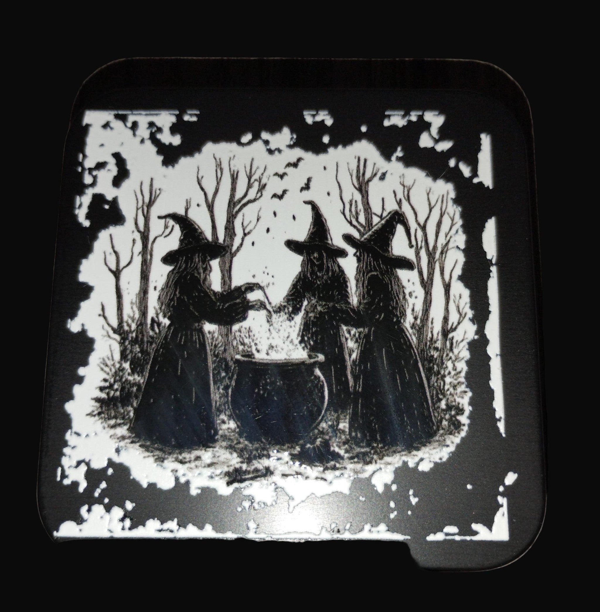 Decorative coaster with black and white illustration of three witches by a cauldron on a wooden surface.