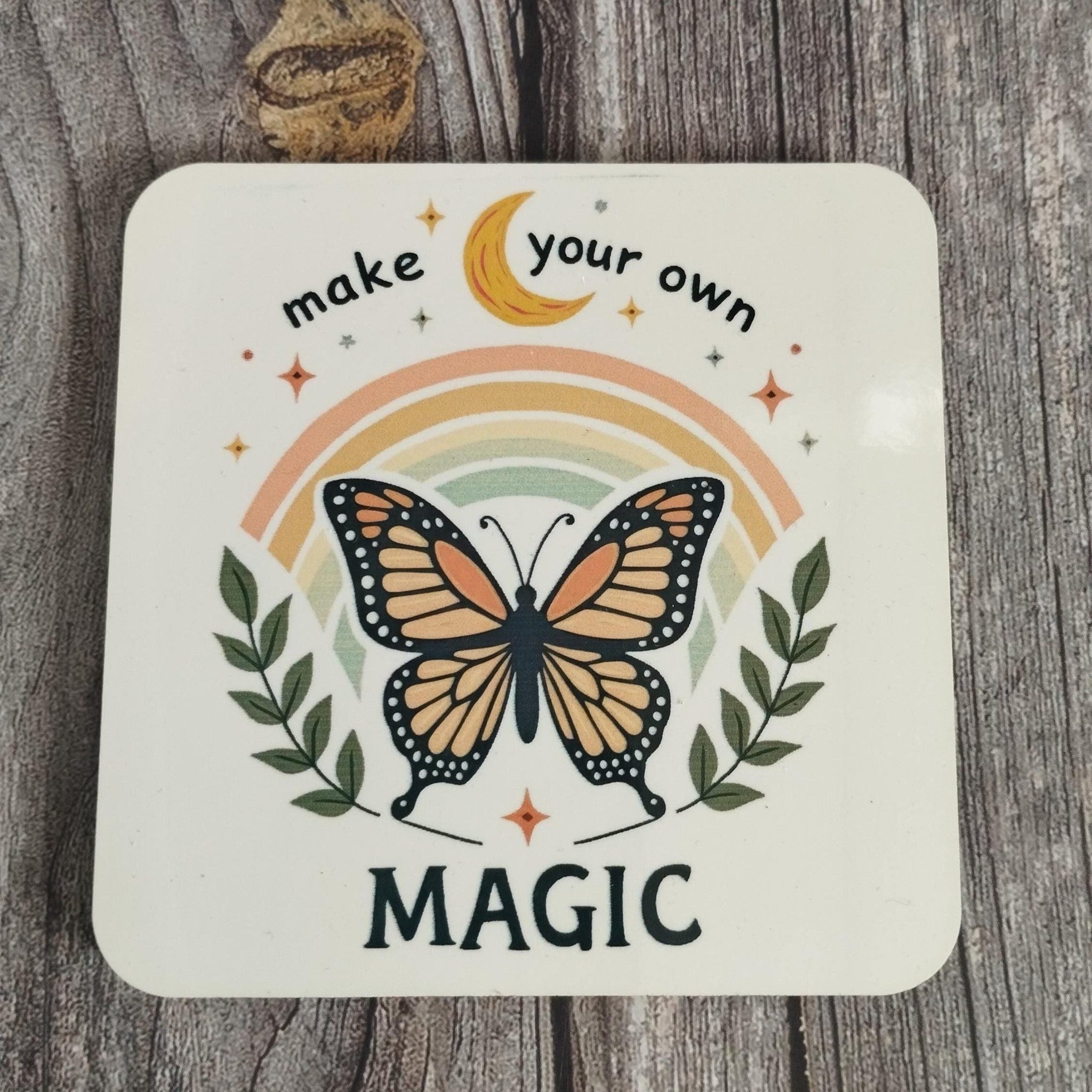 Coasterwith a butterfly, rainbow, and text 'make your own magic' on a wooden surface