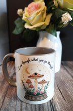 Mug with mushroom design and text on a wooden surface with flowers in the background