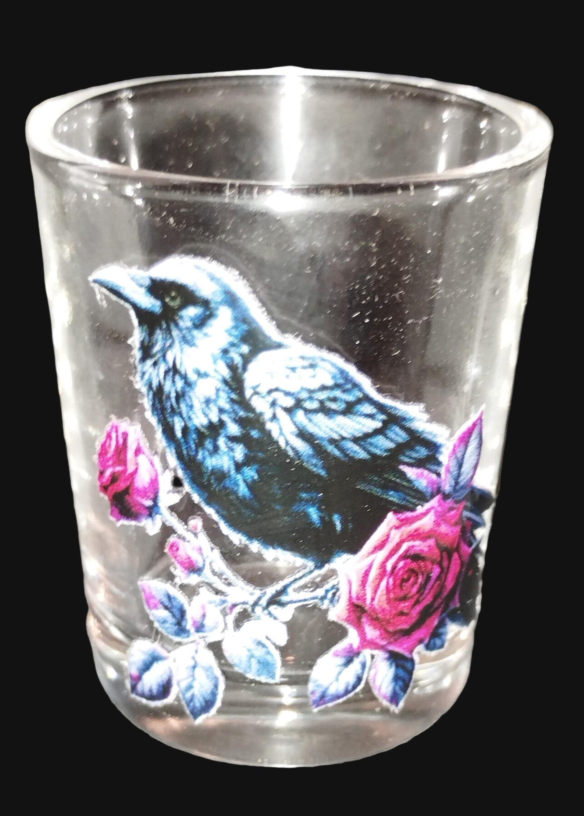 Clear glass shot glass with a raven and pink flowers design on a dark surface.