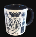 Mug with 'Stay Weird' text and mystical illustrations on a wooden surface.