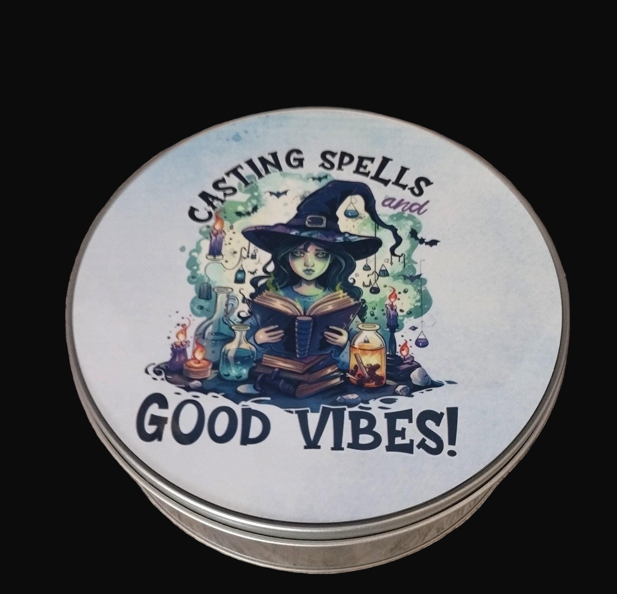 Tin lid with a witch illustration and text 'Casting Spells and Good Vibes!' on a wooden surface.