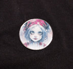 Round fridge magnet showing a pale blue-haired girl in a red winter hat against a snowy pink background