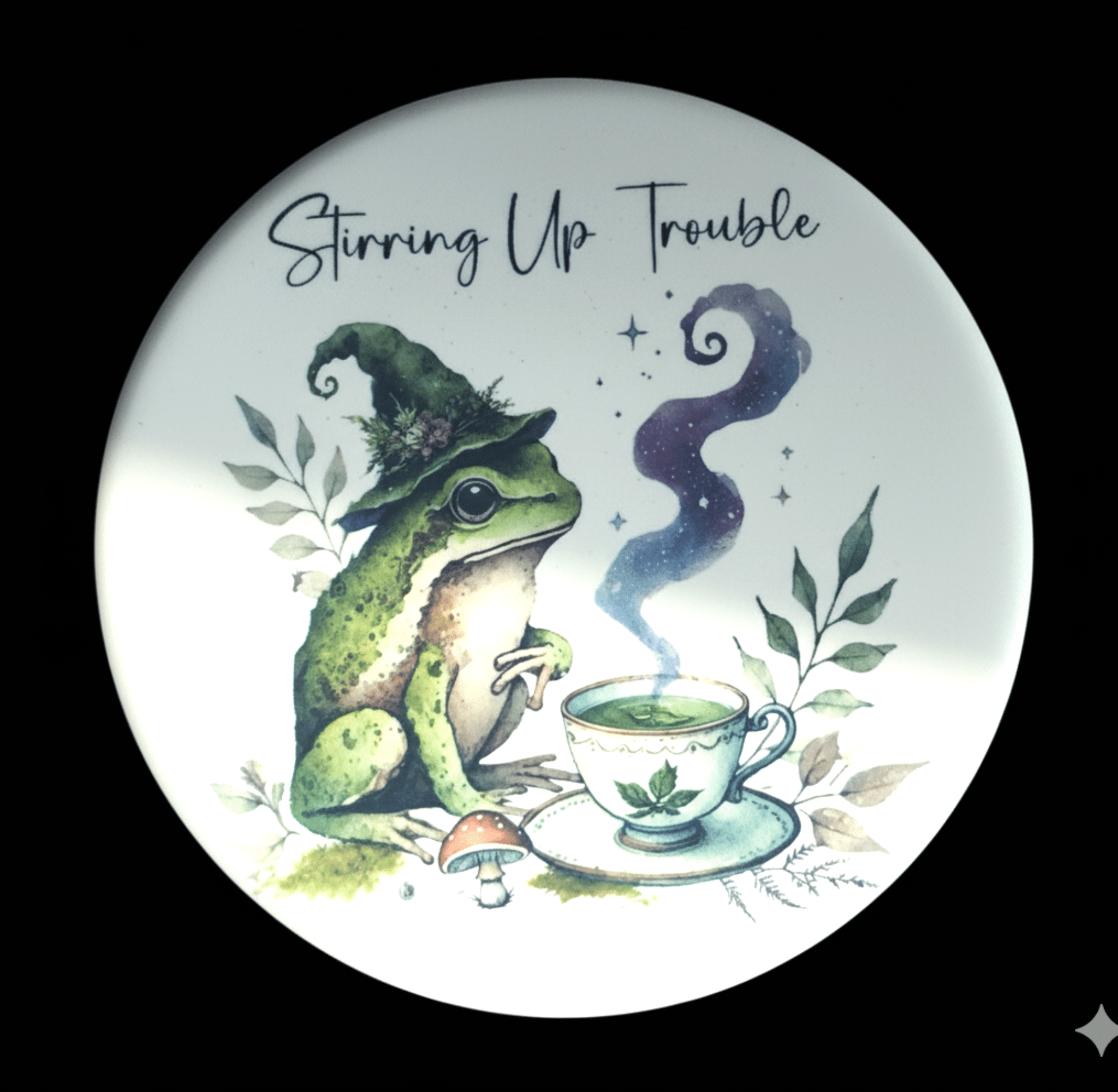 Frog wearing a witch hat with a teacup and text 'Stirring Up Trouble' on a decorative plate.