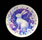 Decorative coaster with a white hare surrounded by flowers and leaves on a black background