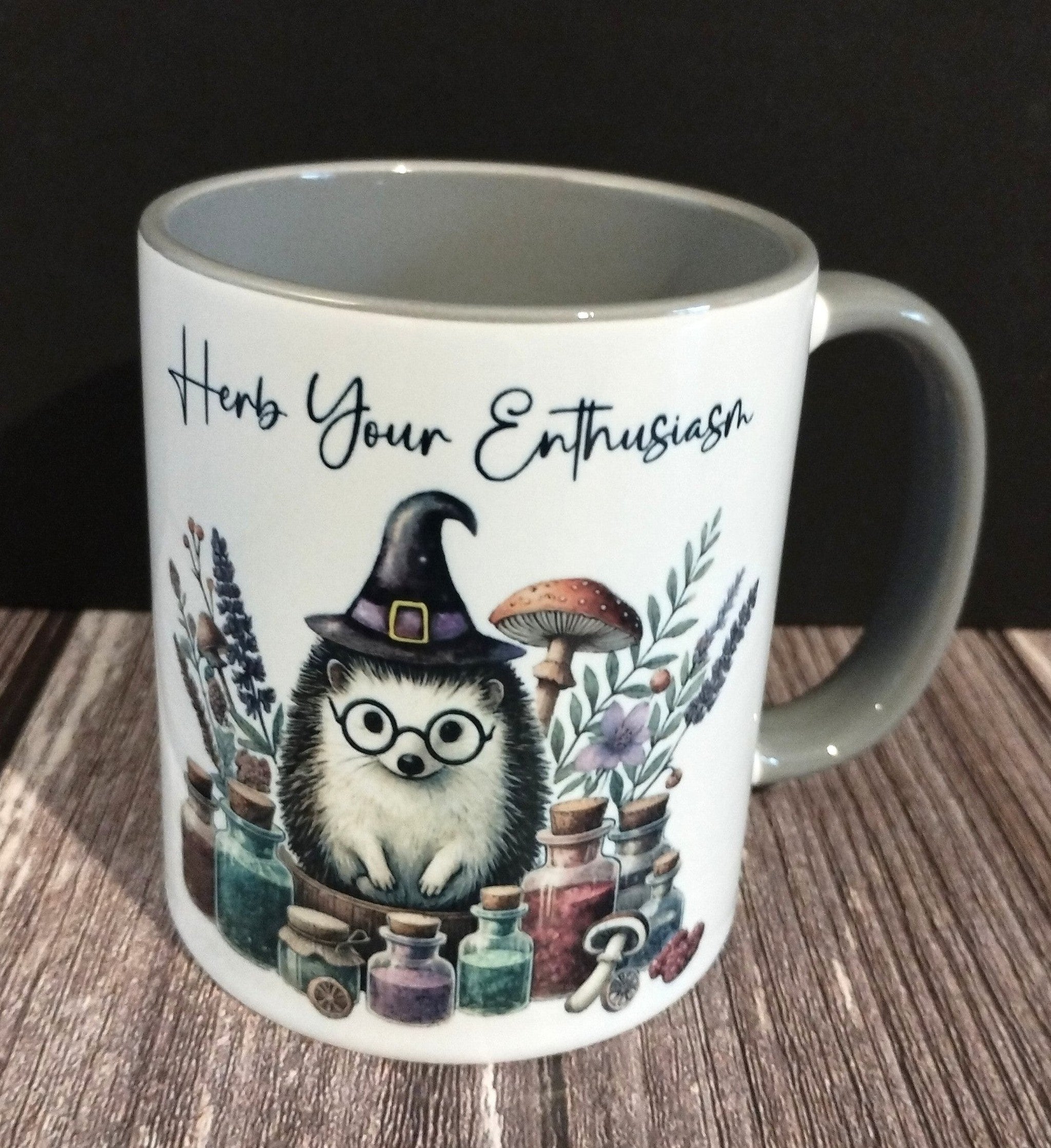 Mug with hedgehog wearing a witch hat and glasses, surrounded by plants and potions, on a wooden surface.