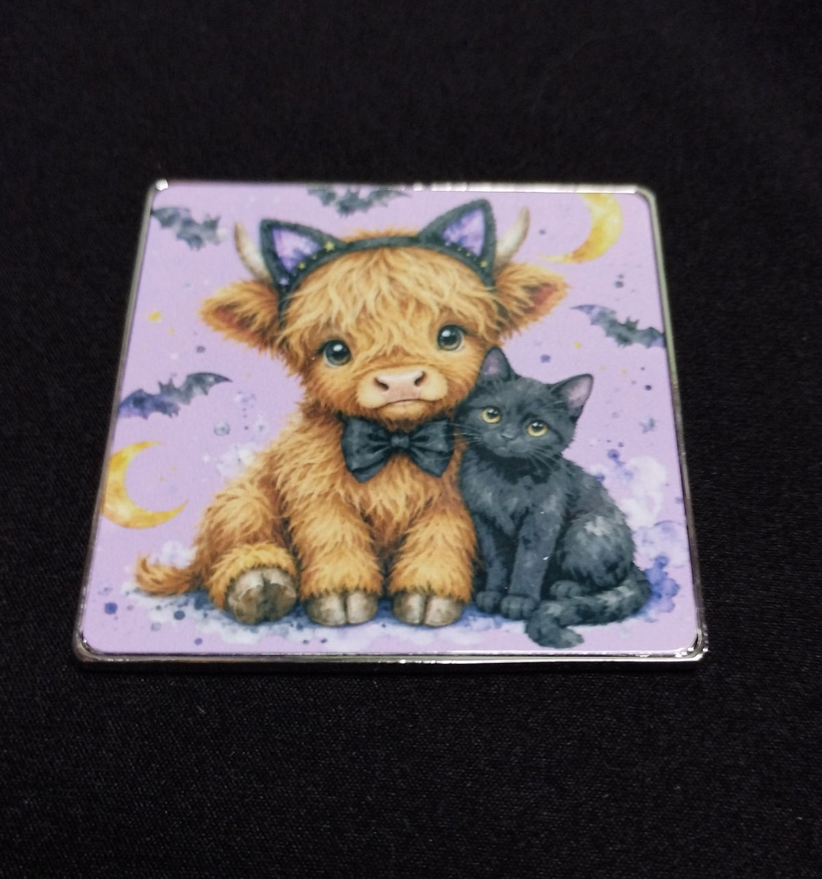 Square Halloween fridge magnet featuring a fluffy Highland cow wearing cat ears and a bow beside a black cat on a lilac background with bats and crescent moons.