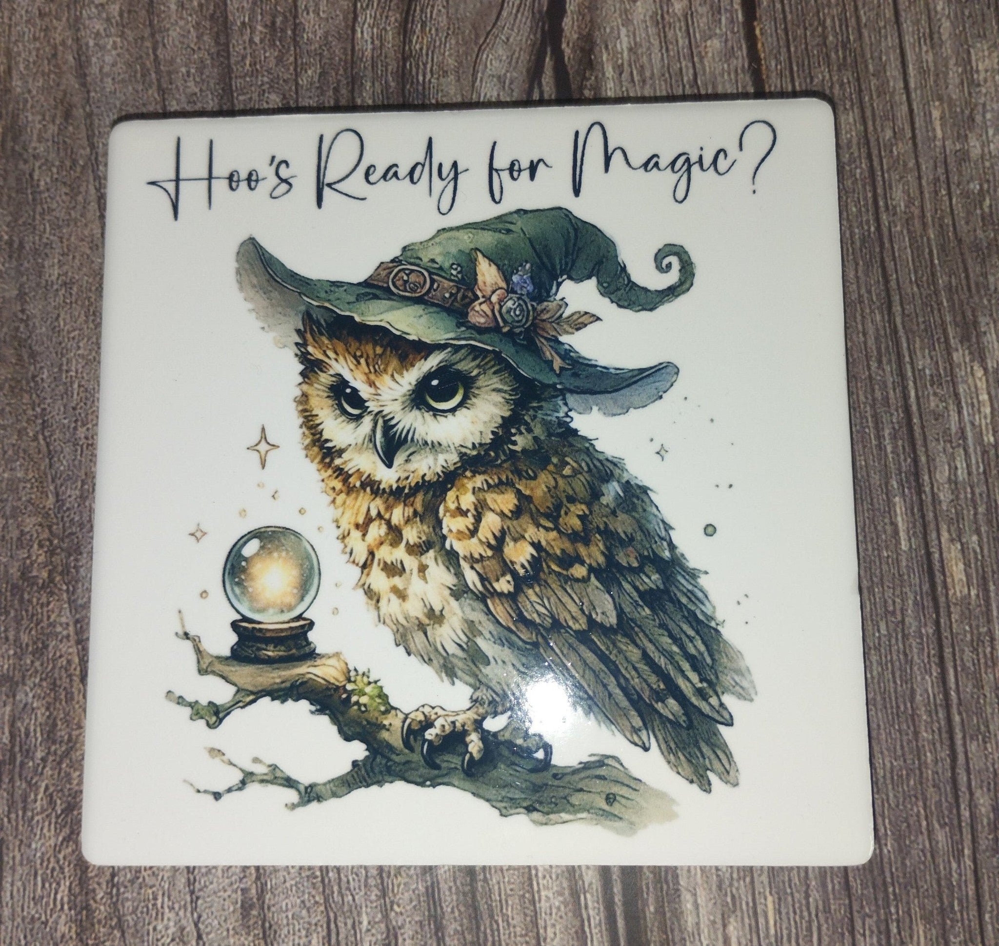 Coaster with an owl wearing a witch hat and crystal ball, text 'Hoo's Ready for Magic?' on a wooden surface.