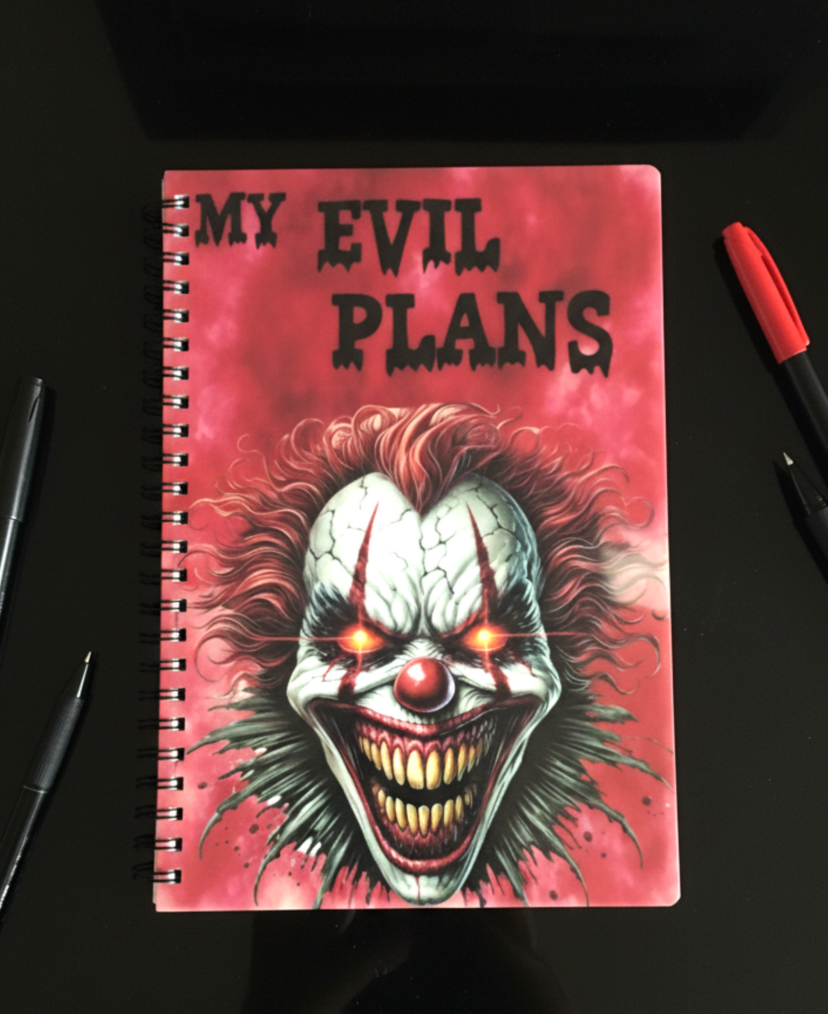 A spiral-bound notebook with a horror-themed cover featuring a creepy clown face and the text 'MY EVIL PLANS' in black on a pink background. The notebook is photographed on a dark surface with pens placed around it.