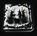 Decorative tin with black and white illustration of three witches and a cauldron on a wooden surface.