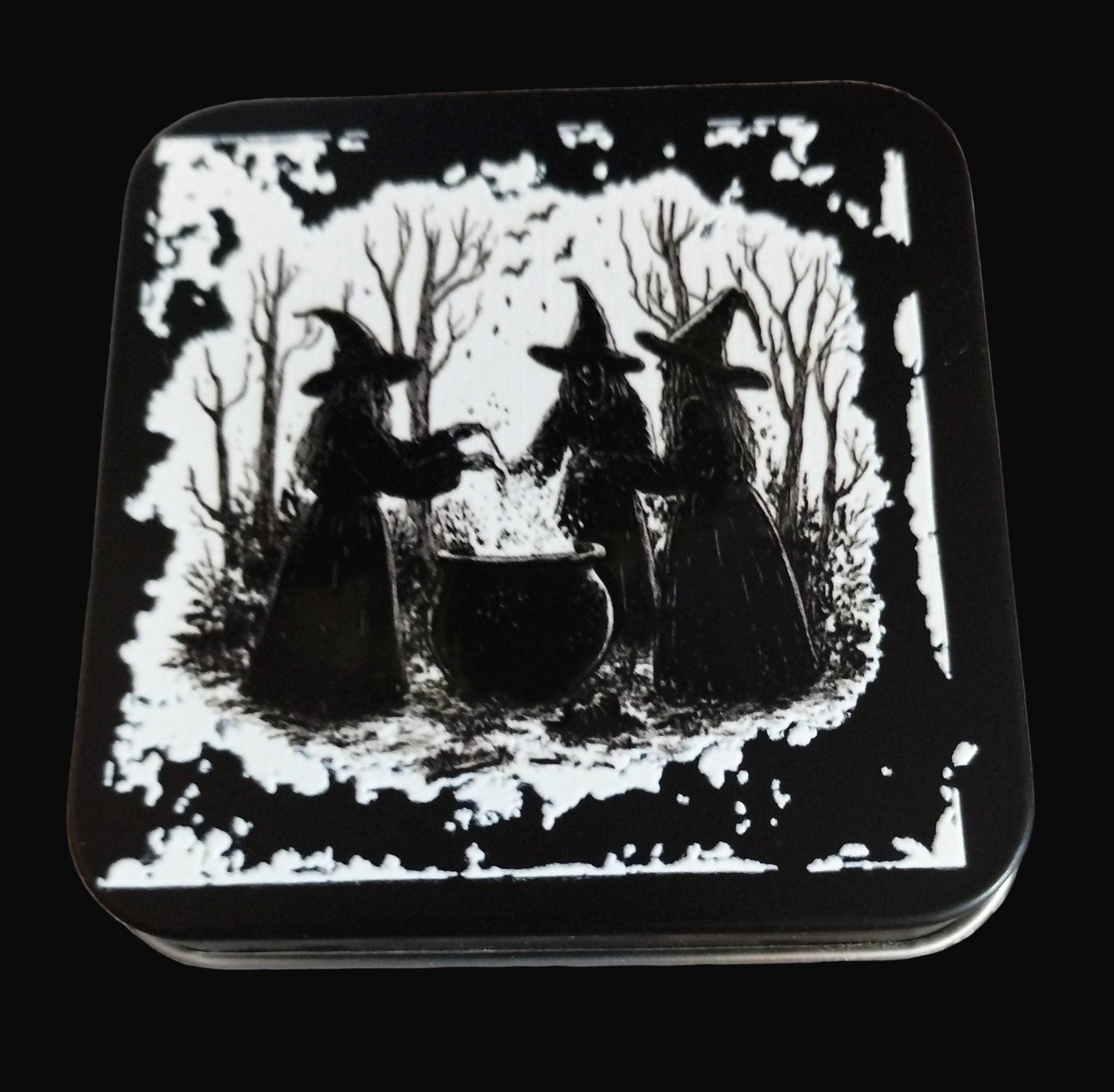 Decorative tin with black and white illustration of three witches and a cauldron on a wooden surface.