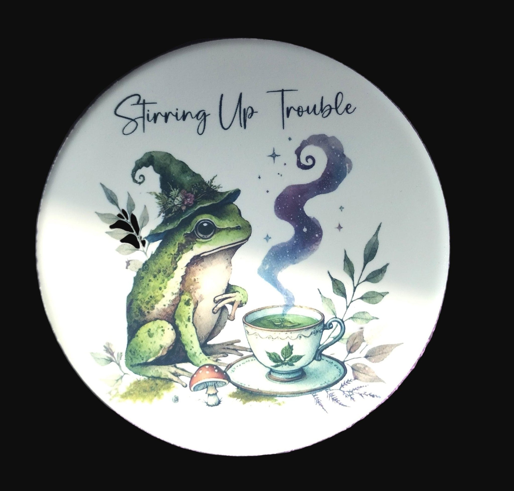 Round coaster with a frog wearing a witch hat and brewing tea, text 'Stirring Up Trouble' on a purple textured background.