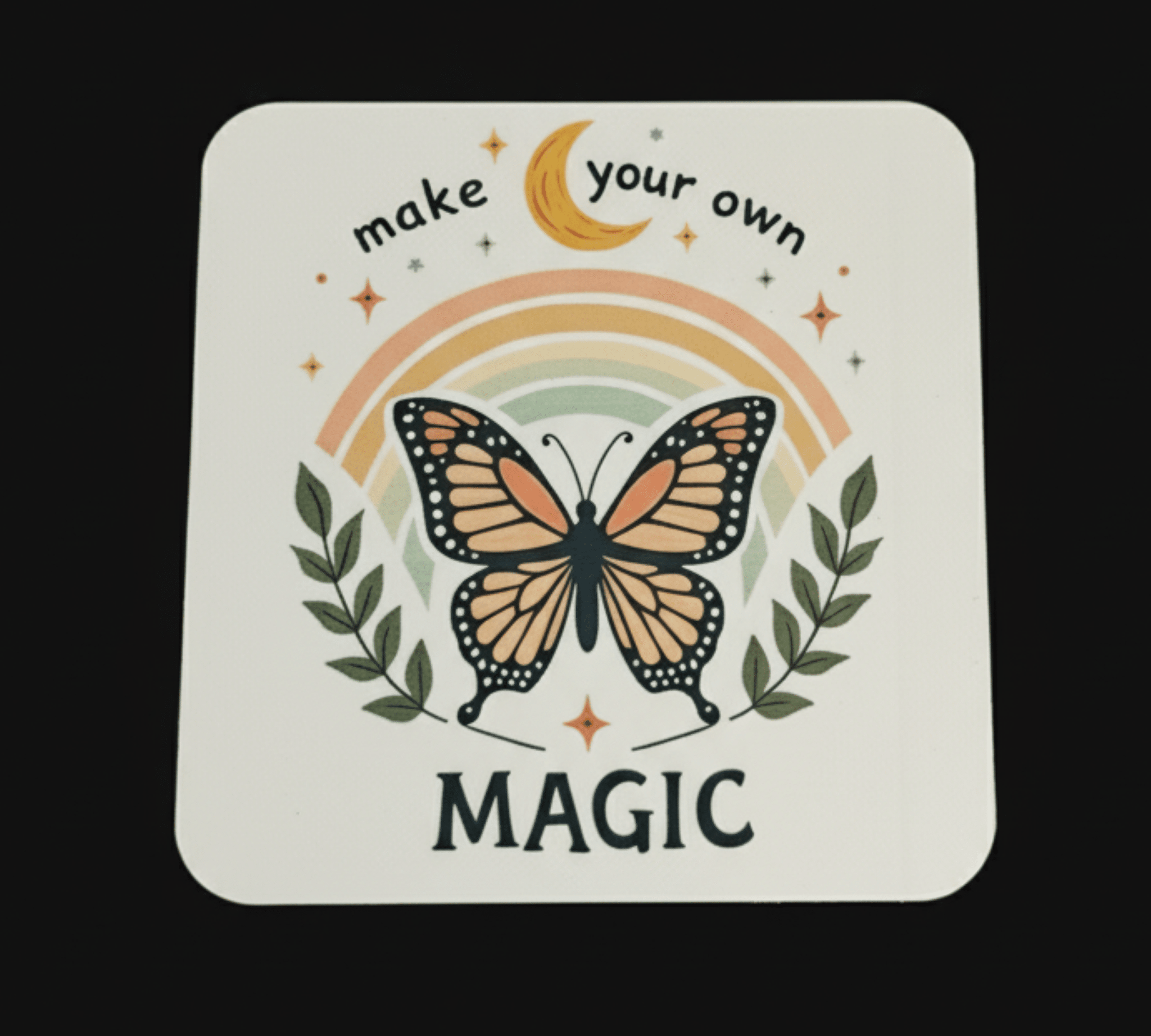 Coaster with butterfly design, rainbow, and text 'make your own magic' on a black background