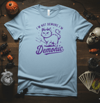 Light blue t-shirt with purple graphic and text on a wooden surface with Halloween decorations.