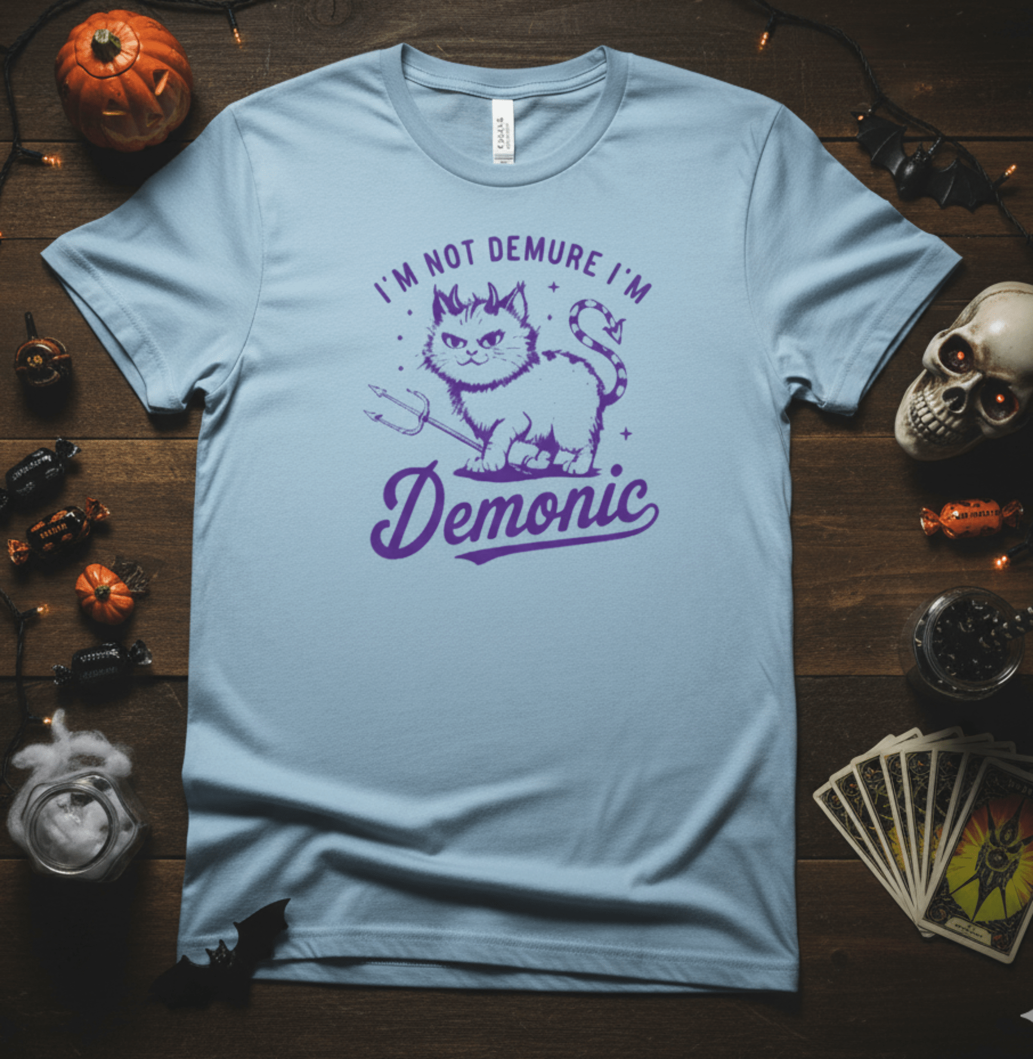 Light blue t-shirt with purple graphic and text on a wooden surface with Halloween decorations.