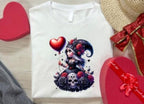 White t-shirt with a colorful graphic design featuring a character with a heart, surrounded by roses and skulls, on a wooden surface with decorative items.