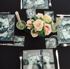 Table setting with floral centerpiece, 'Witch's Brew' themed coasters, and glasses on a black surface.