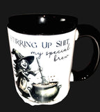 Mug with witch design and text 'Stirring up shit, my special brew' on a wooden surface.