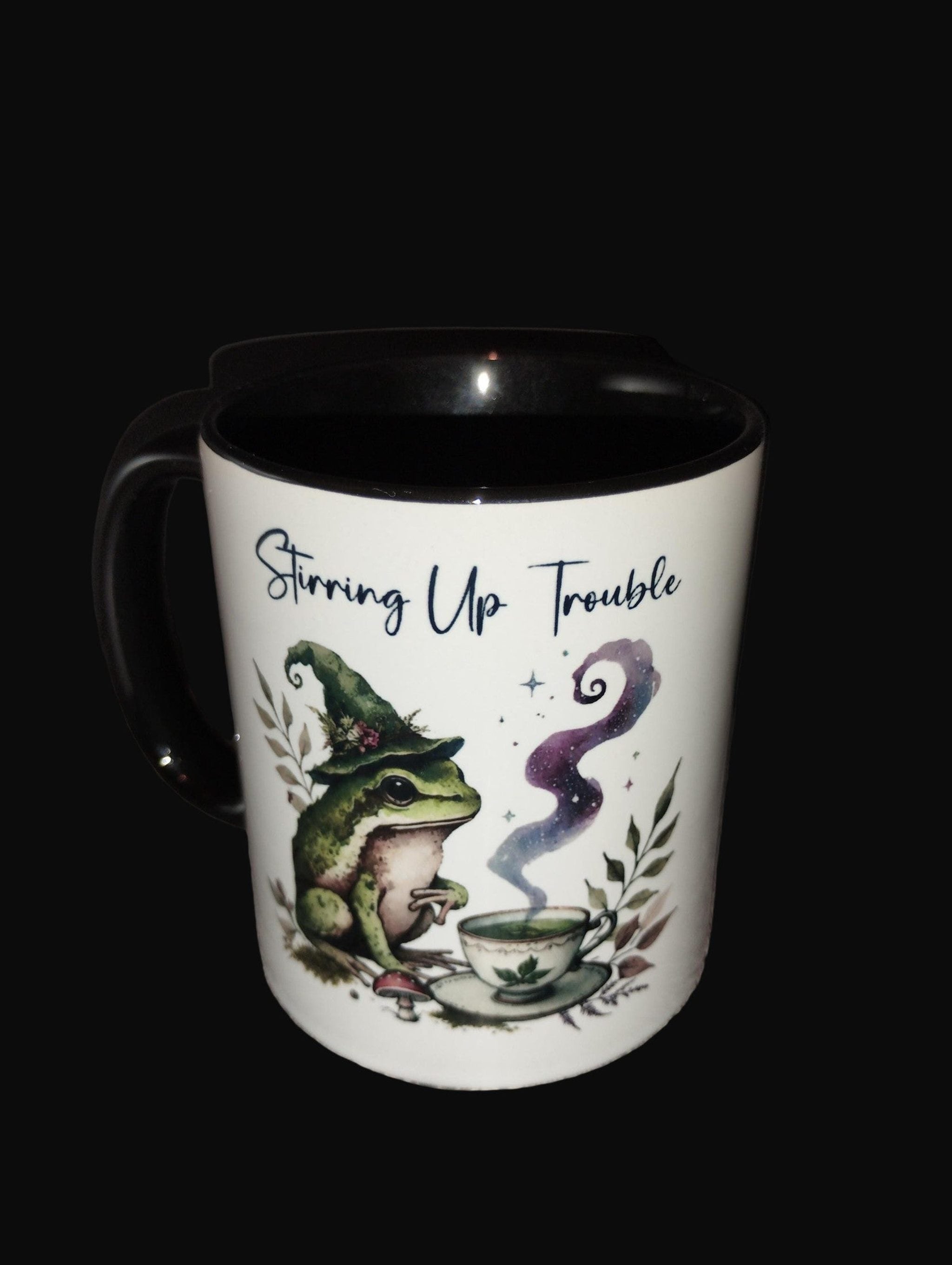 Mug with a frog wearing a witch hat and text 'Stirring Up Trouble' on a wooden surface.