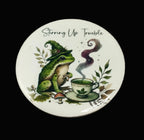 A round white ceramic plate or coaster featuring a whimsical illustration of a green frog in a witch hat next to a steaming teacup, with the text 'Stirring Up Trouble' above. The item is photographed flat on a rustic, dark wood-textured background.