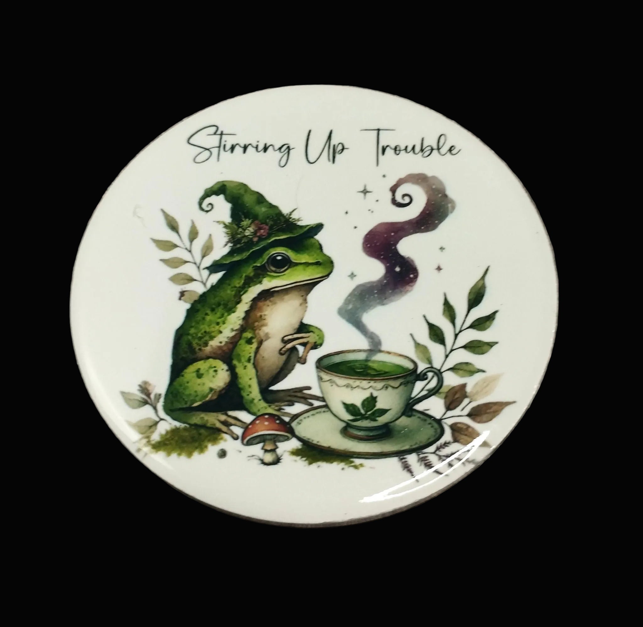A round white ceramic plate or coaster featuring a whimsical illustration of a green frog in a witch hat next to a steaming teacup, with the text 'Stirring Up Trouble' above. The item is photographed flat on a rustic, dark wood-textured background.