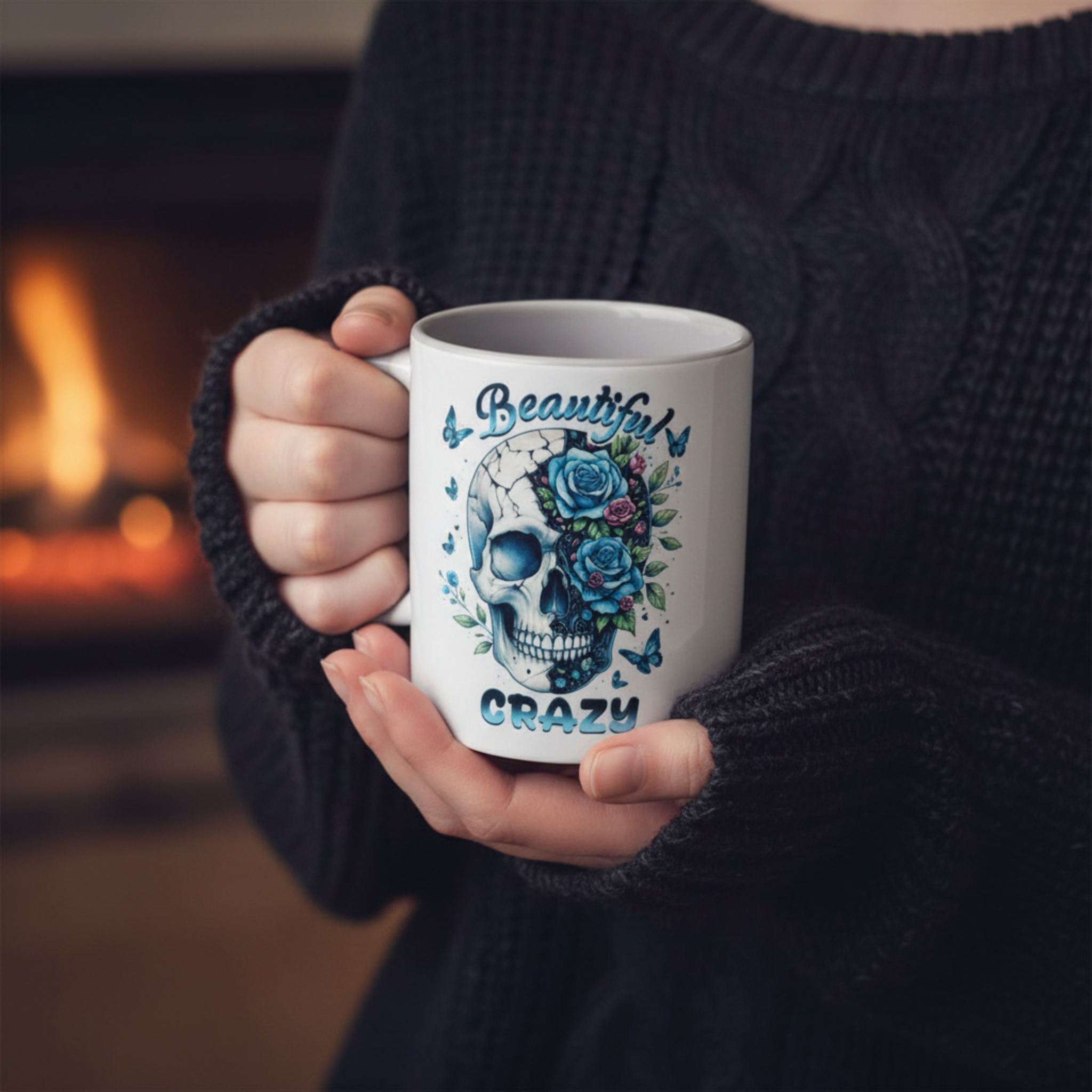 A person in a dark cable-knit sweater holds a white ceramic mug with a blue and black skull graphic decorated with roses and butterflies, with the words 'Beautiful CRAZY' printed on it. The background shows a warm, blurred fireplace.