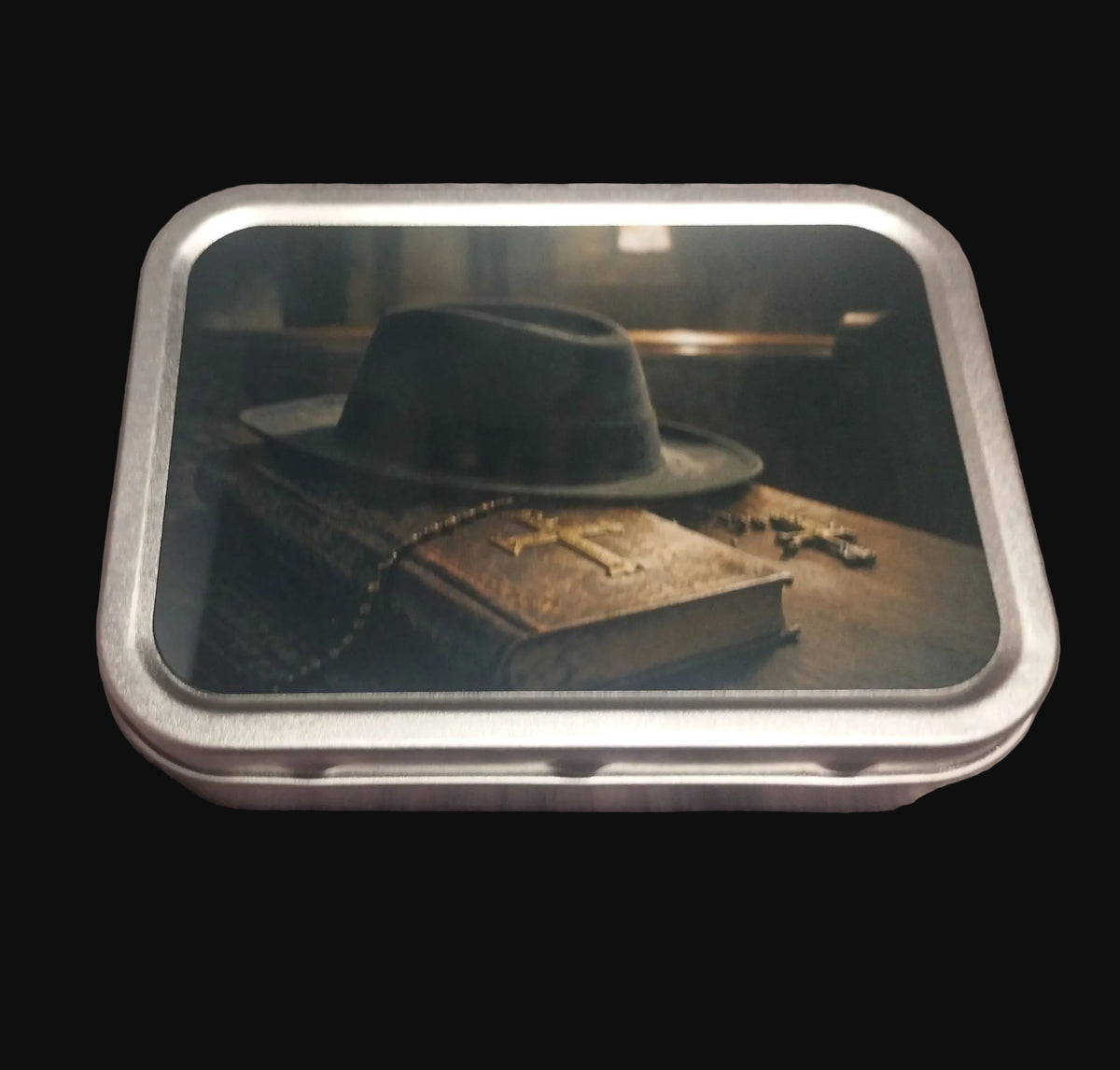 Silver-coloured rectangular tobacco tin with dark artwork showing a black hat on an aged book with cross and rosary-style details.
